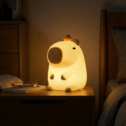 A glowing squishy silicone capybara night light with a small orange on its head, sitting on a wooden bedside table next to an open book and glasses in a warm, dimly lit bedroom.
