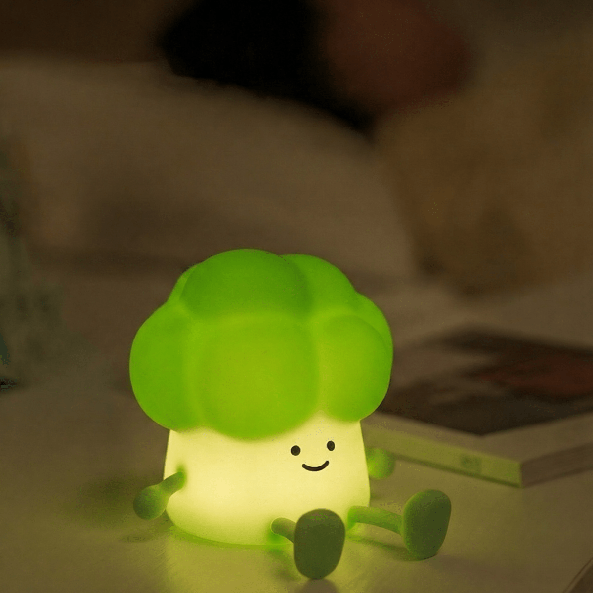 A cute smiling broccoli squishy silicone night light emitting a warm green glow on a white bedside table next to an open book in a dimly lit, cozy bedroom.