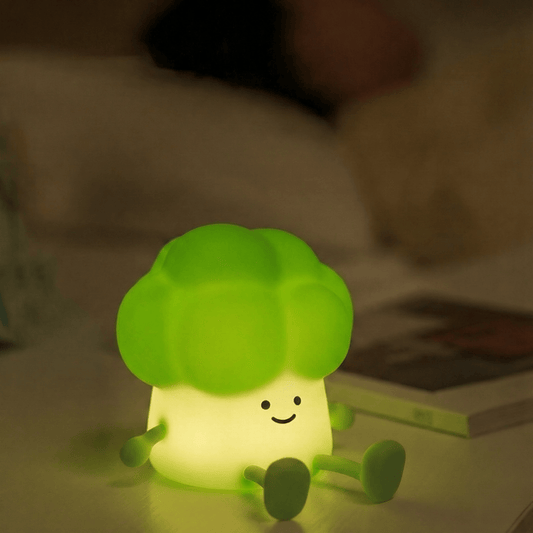 A cute smiling broccoli squishy silicone night light emitting a warm green glow on a white bedside table next to an open book in a dimly lit, cozy bedroom.