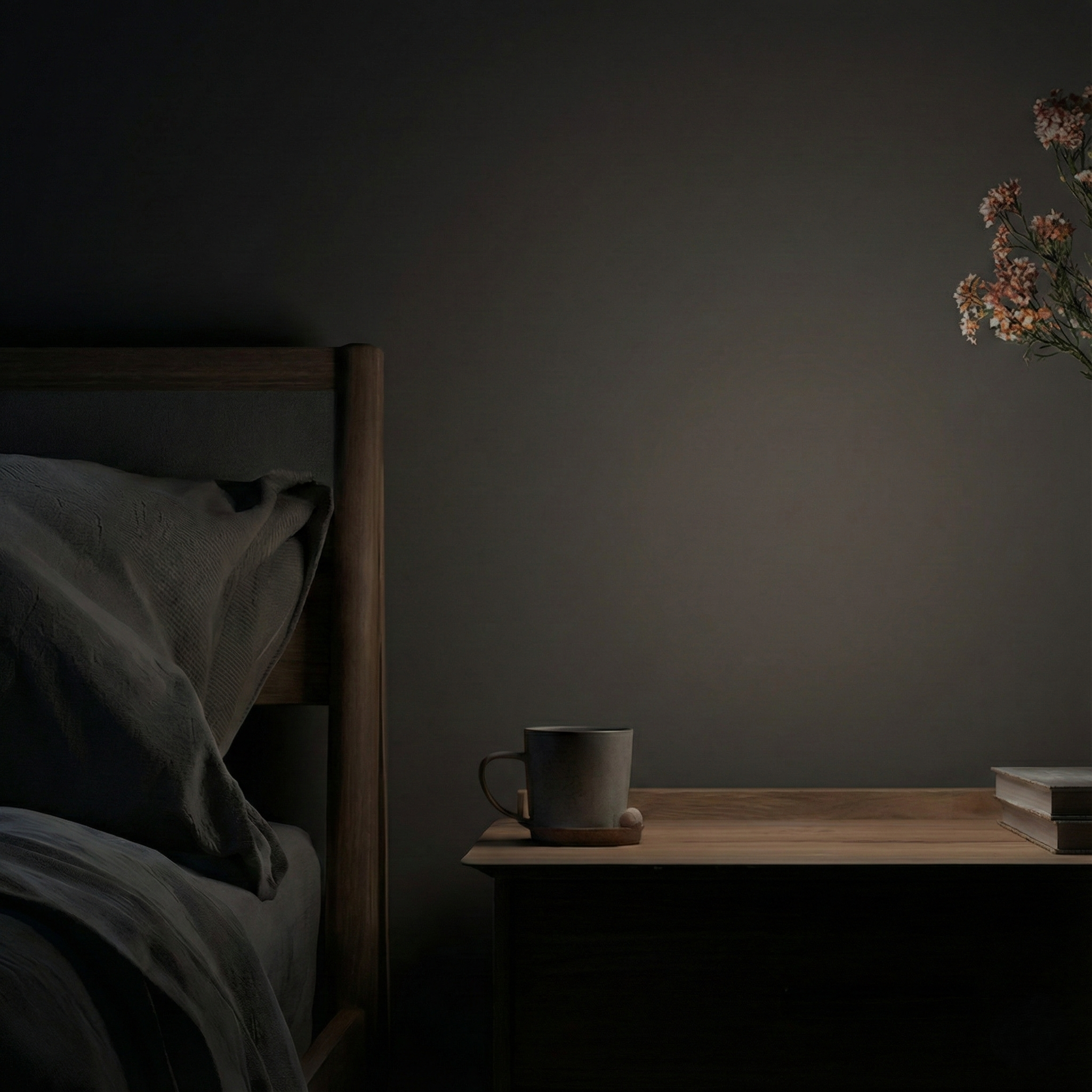 A dark and moody minimalist bedroom scene featuring a wooden nightstand with a grey mug and books, next to a cozy bed with dark grey pillows against a charcoal wall.