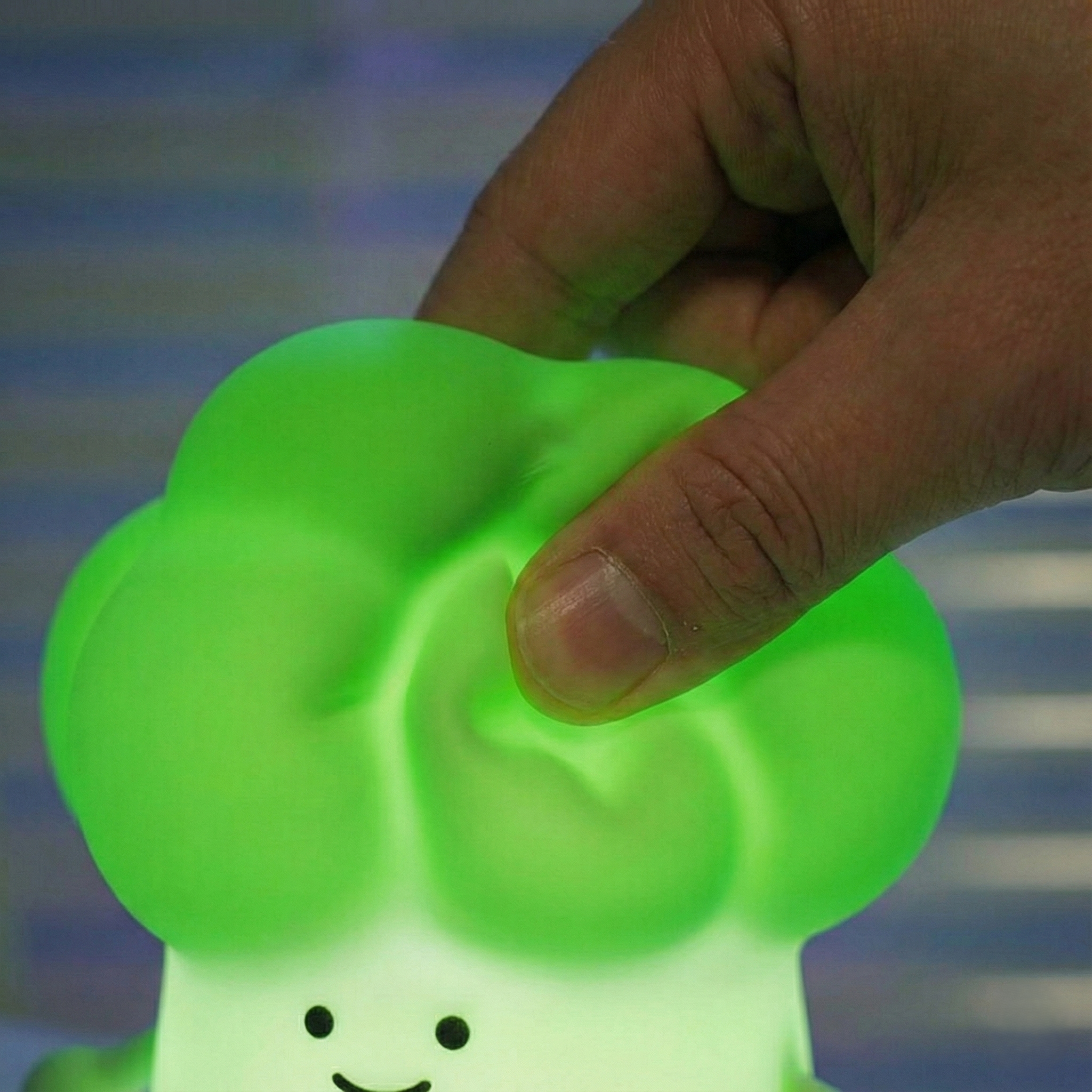 Close-up of a hand squeezing a glowing green broccoli silicone night light with a cute smiley face, showing its soft and flexible material.
