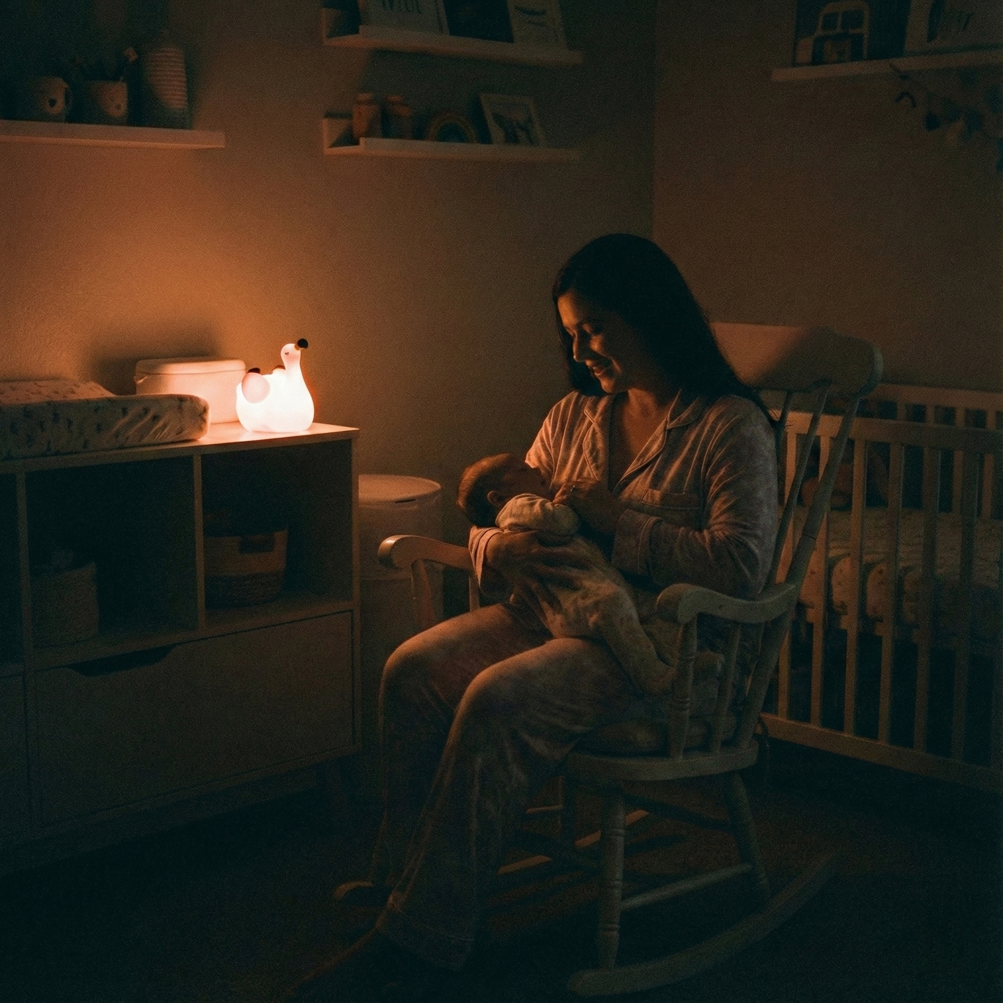 A mother sitting in a wooden rocking chair, cradling her baby in a cozy, dimly lit nursery. A glowing Squiloo flamingo night light sits on a changing table nearby, casting a warm, comforting orange light.