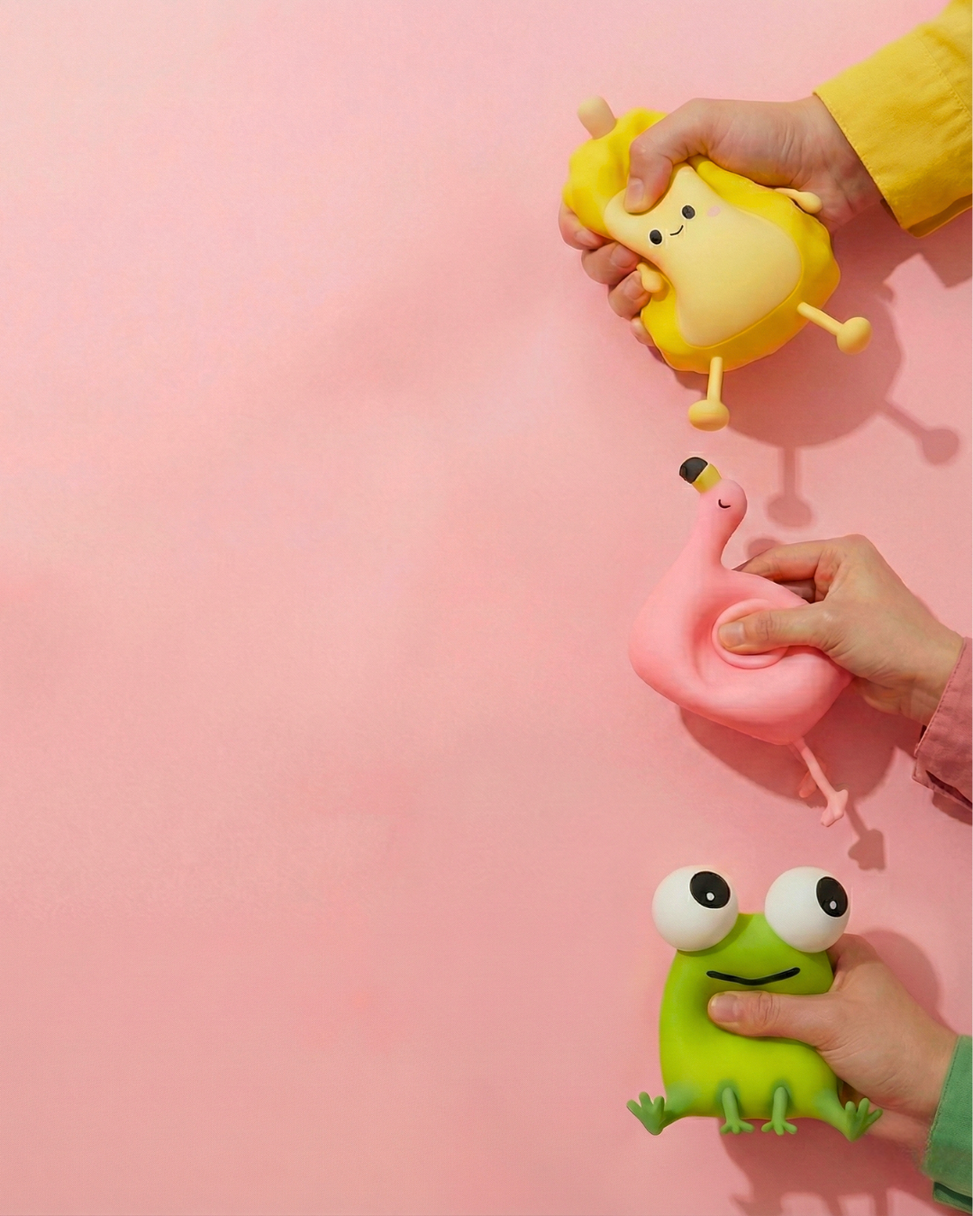 Three hands squeezing three different colorful squishy silicone night lights—a yellow mango, a pink flamingo, and a green frog—against a vibrant pink background to demonstrate their soft, anti-stress texture.