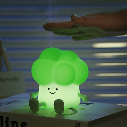 Glowing green squishy broccoli silicone night light with a happy face sitting on a book, with a person's hand hovering above it to demonstrate tap-control activation.