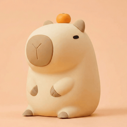 Cute squishy silicone capybara night light with a small orange on its head, sitting against a soft peach-colored background.