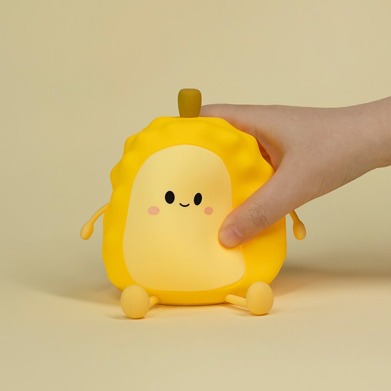 Cute yellow squishy durian silicone night light with a happy face sitting on a dark sofa next to yellow building blocks and a book.