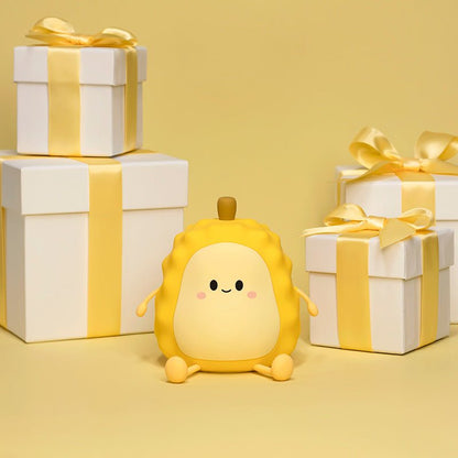 Cute yellow squishy durian silicone night light with a happy face sitting on a dark sofa next to yellow building blocks and a book.