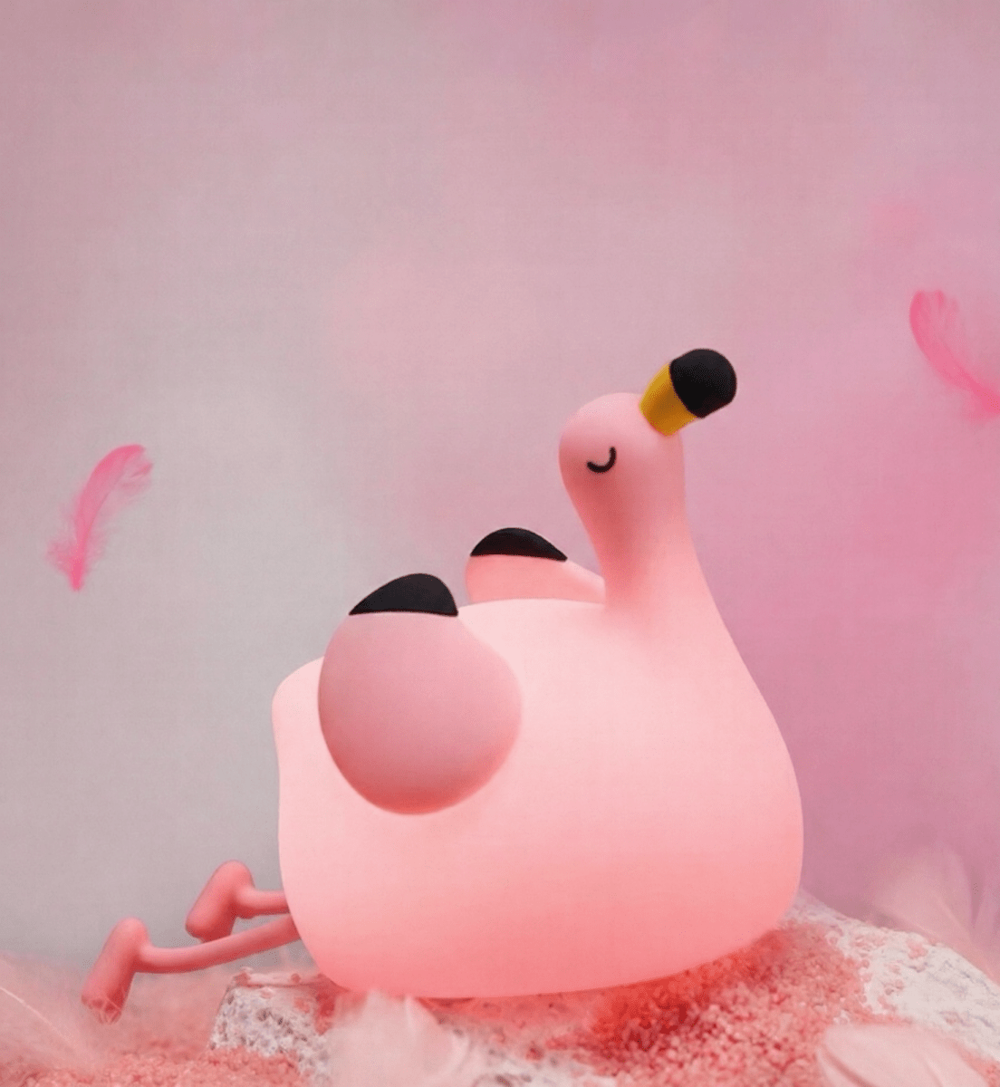 Dreamy pink squishy flamingo silicone lamp lying on a soft pink surface surrounded by floating pink feathers in a magical, aesthetic setting.