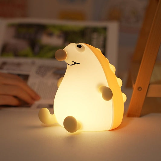 Cute yellow and white squishy silicone hedgehog night light with a happy face, glowing warmly on a white desk with a cozy blurred background.