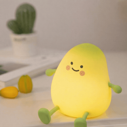 Cute yellow squishy mango silicone night light with a happy face and little limbs glowing on a white surface with a small cactus in the background.