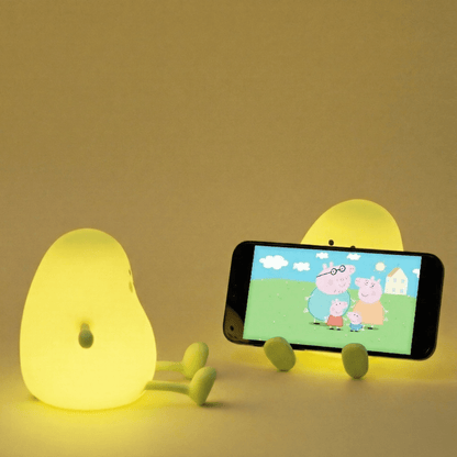 Two glowing yellow mango squishy silicone lamps on a beige background, with one lamp holding a smartphone horizontally displaying a cartoon.