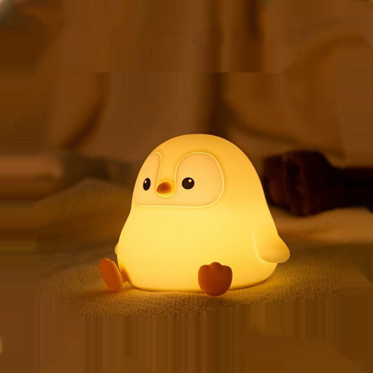 Cute yellow squishy penguin silicone lamp sitting on a soft surface, emitting a warm golden glow in a dimly lit room.