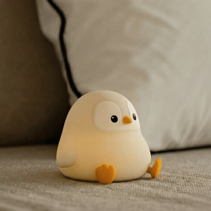 Cute white squishy penguin silicone lamp with orange feet sitting on a textured grey surface and glowing warmly against a soft blurred background with a pillow.